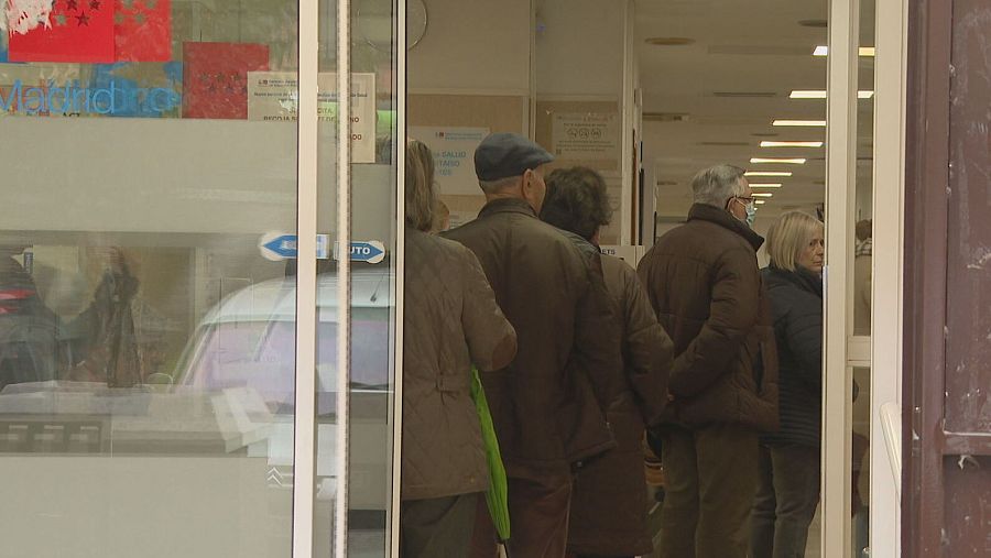 Personas esperando en fila ante una puerta de cristal de un centro sanitario.  Una flecha indica la entrada. Se observan carteles en la puerta.