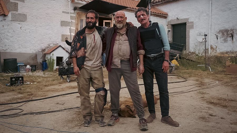 Tres actores posan en un set de rodaje exterior, con vestuario variado y un perro presente.  Se aprecian elementos de utilería y equipo de filmación en un entorno rural.