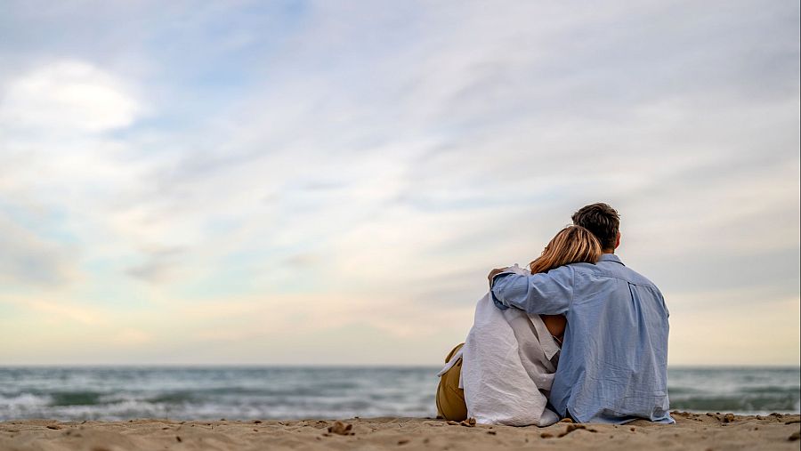 Pareja abrazada en una playa con cielo nublado y mar tranquilo; la mujer lleva una manta.