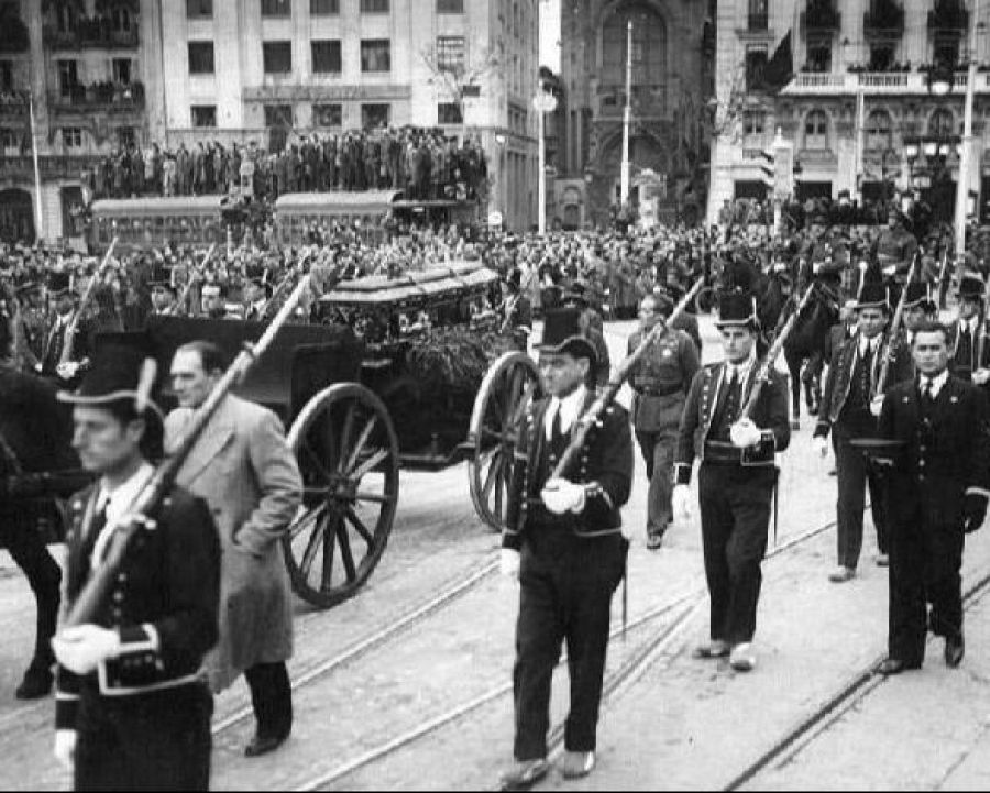 Cortejo fúnebre en blanco y negro: carruaje con féretro cubierto, escoltado por agentes uniformados y rodeado de multitud.