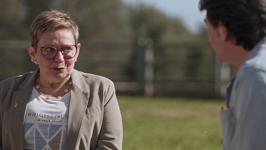 Mujer con camiseta "BRIGHTNESS IN YOUR HEART" y gafas habla con un hombre en un entorno exterior con árboles y valla.