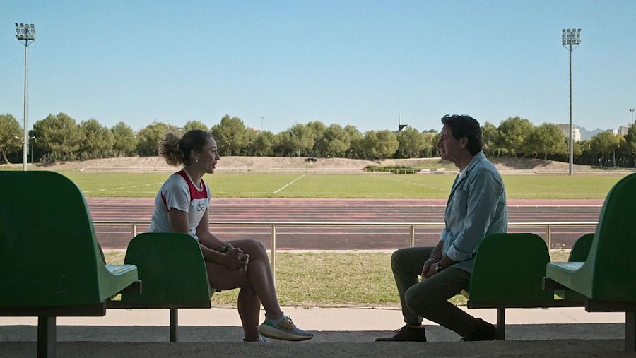 Conversación distendida entre una mujer joven con camiseta deportiva y un hombre mayor en las gradas de una pista de atletismo, en un día soleado.