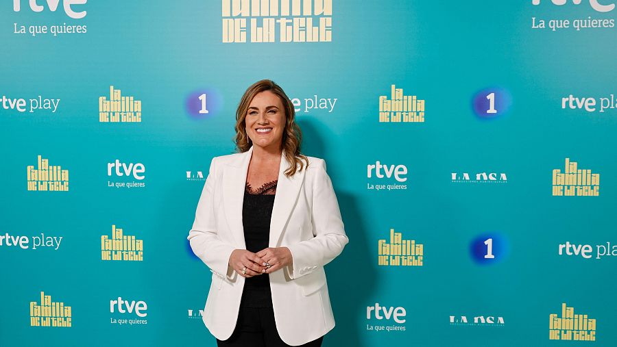 Presentadora de televisión sonriendo ante un photocall turquesa con logos de RTVE y el título del programa en letras doradas.
