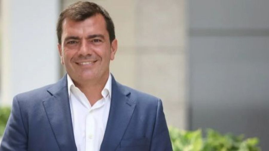 Retrato de un hombre de mediana edad, sonriente, con traje azul marino y camisa blanca. Fondo urbano desenfocado.