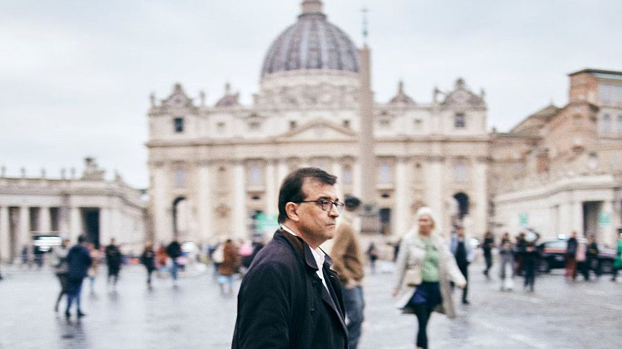 Hombre con gafas y abrigo oscuro en la Plaza de San Pedro, mirando hacia la derecha con expresión seria.  Al fondo, la Basílica de San Pedro.