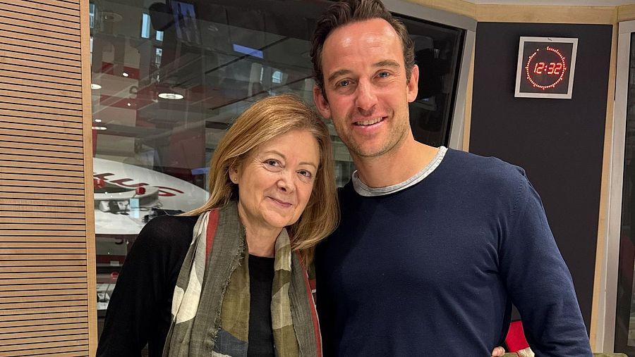 Foto de dos personas de mediana edad en un estudio de radio. Ella, con cabello rubio y bufanda; él, con cabello castaño y jersey azul.  Reloj digital marcando las 12:32.