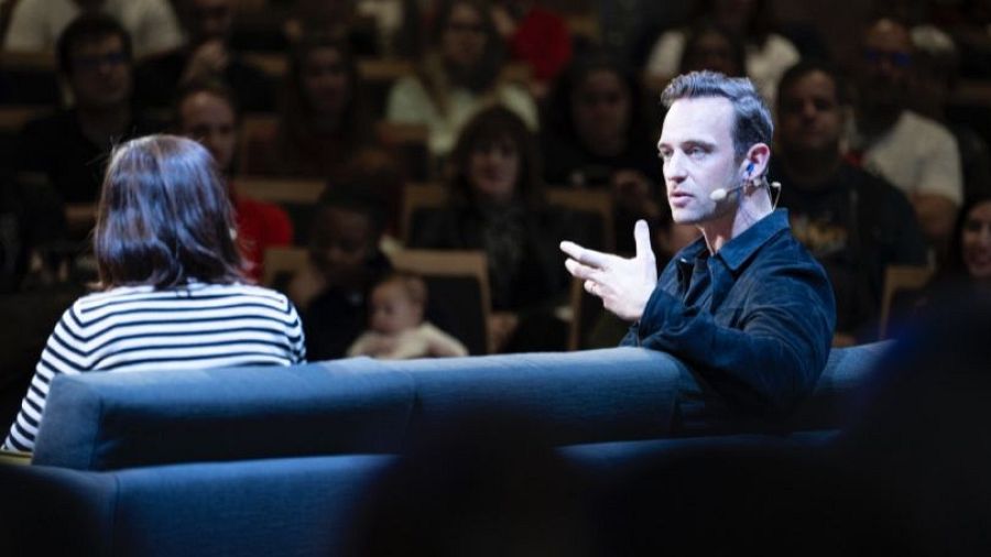 Entrevista o charla con Eduardo Casanova, actor y director, quien gesticula con un micrófono de diadema mientras está sentado en un sofá junto a una mujer.