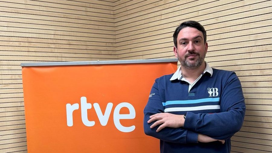 Piloto de sumergibles en entrevista de radio, con polo azul marino a rayas y brazos cruzados, en estudio con panel de madera y roll-up naranja con logo RTVE.
