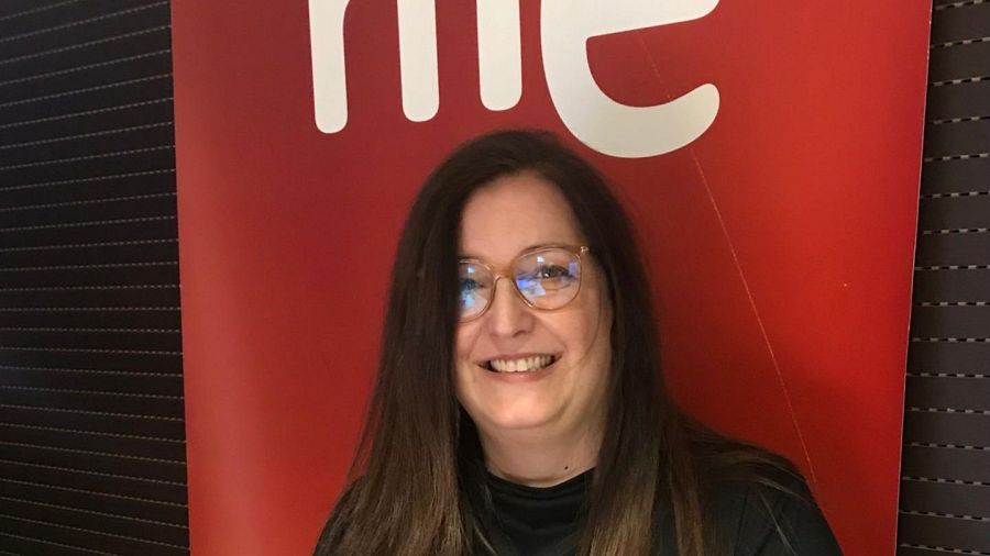 Mujer con gafas sonríe ante fondo rojo con la palabra "rine" y marrón oscuro con textura de madera. Lleva ropa oscura.