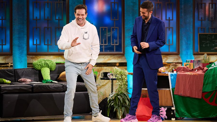 Actuación cómica en plató de TV con decoración asiática. Un hombre en sudadera blanca gesticula, mientras otro en traje azul lo observa.  Sofá con cojines vegetales y objetos decorativos en la mesa.