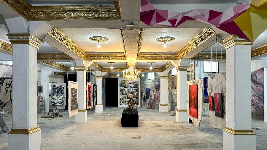 Galería de arte con techo alto, columnas blancas y doradas, paredes de ladrillo visto y pintura blanca, exponiendo pinturas, collages y otras obras de arte.