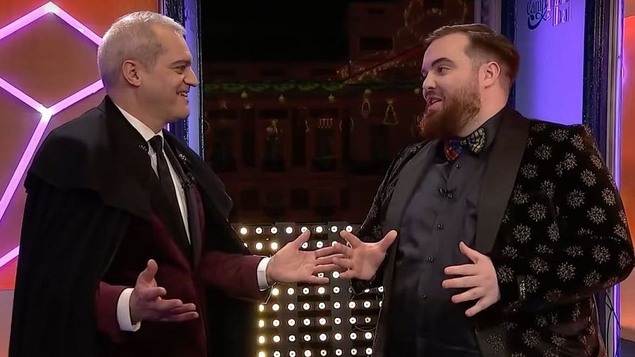 Ibai Llanos y Ramón García conversan en un plató de televisión; él viste americana negra con detalles brillantes, y él, capa negra sobre traje.