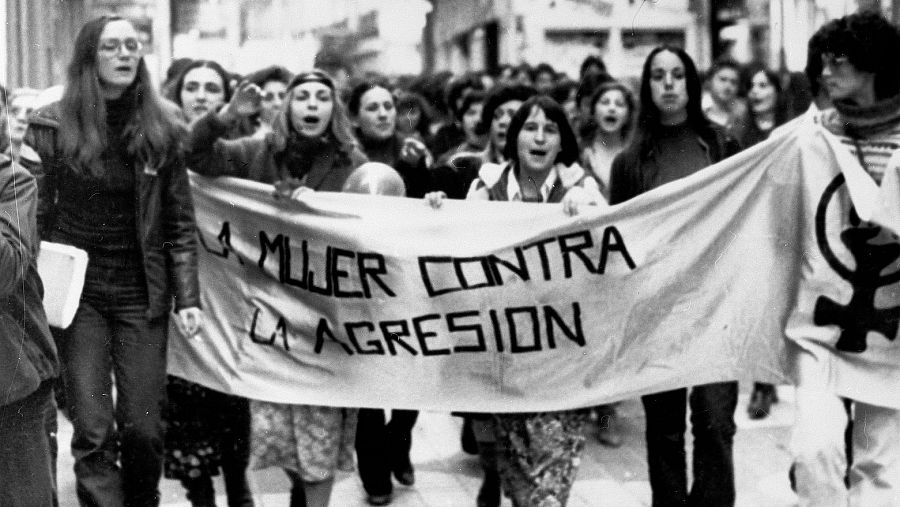 Manifestación de mujeres con pancarta "LA MUJER CONTRA LA AGRESIÓN".  Imagen en blanco y negro; las mujeres muestran expresiones decididas.