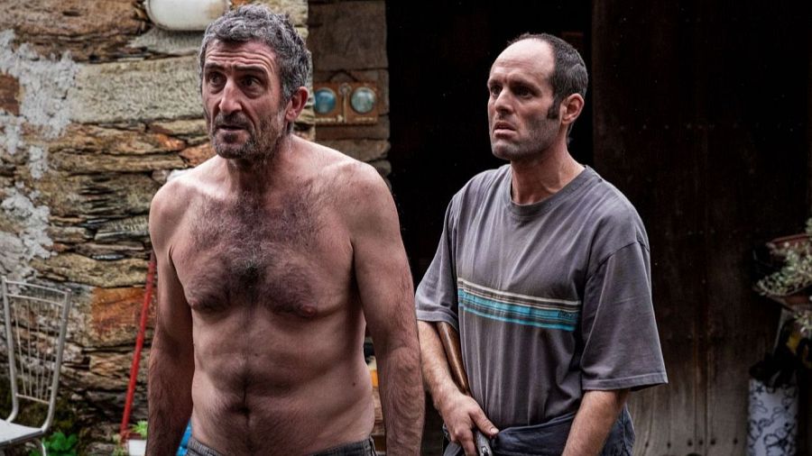 Escena de 'As bestas': dos hombres, uno sin camisa y otro con camiseta gris, con expresiones serias en entorno rural. Uno parece sujetar algo.