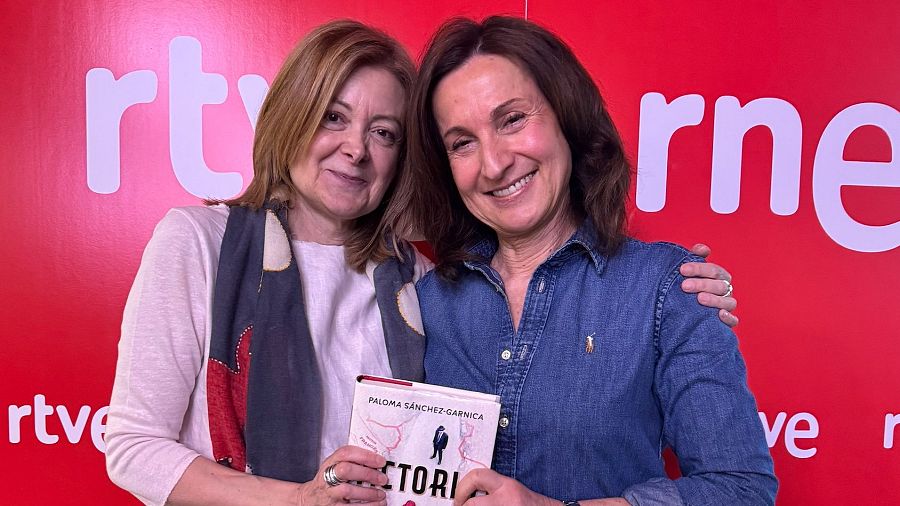 Dos mujeres sonrientes presentan el libro "Victoria" ante un fondo rojo con el logo de RTVE. Una lleva camisa blanca y pañuelo; la otra, camisa vaquera.