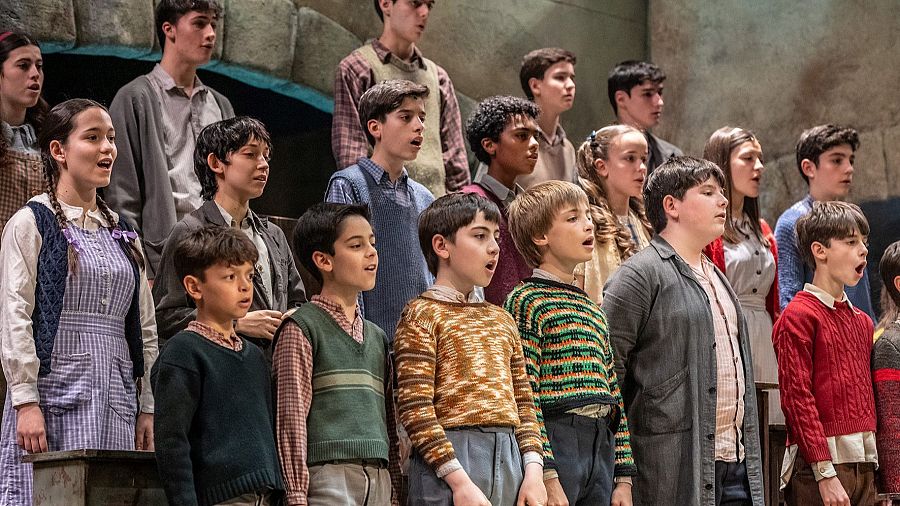 Coro infantil cantando "ESA DIVA" en escenario con pared de piedra; vestuario de época; posible ensayo o actuación.
