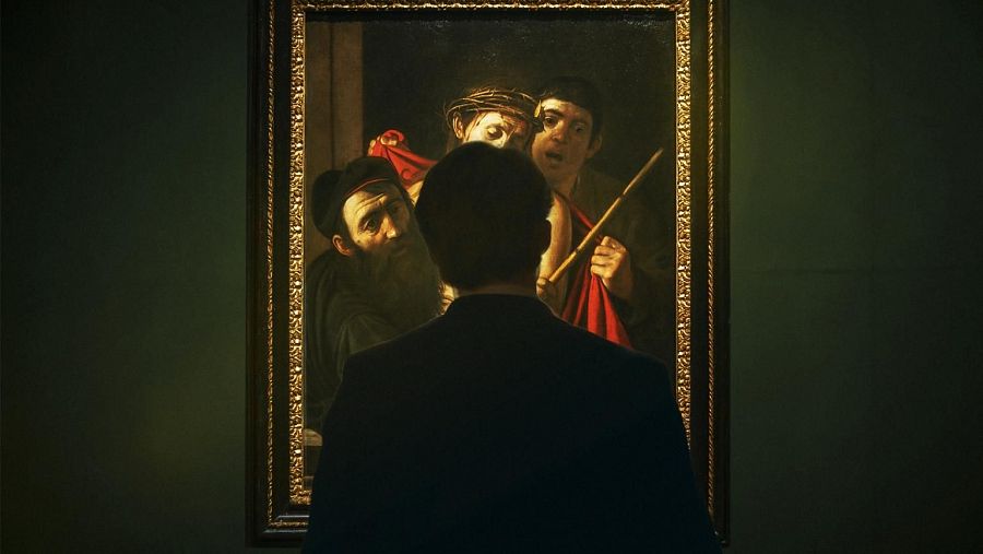 Persona contemplando un cuadro con marco dorado; escena religiosa con tres figuras, iluminación tenue y fondo oscuro.