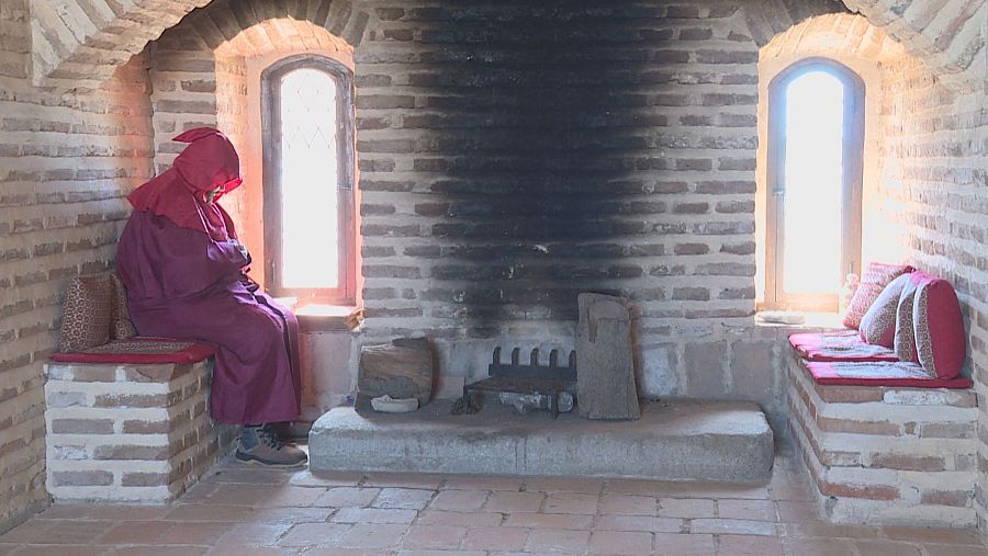 Habitación histórica con chimenea, bancos con cojines y un figurante con túnica granate sentado junto a una ventana arqueada.