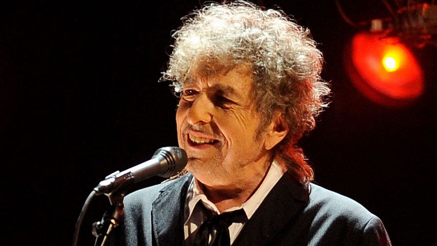 Imagen en blanco y negro de Bob Dylan en concierto.  Viste traje oscuro y está frente a un micrófono, con expresión serena. Iluminación tenue con luz roja difusa.