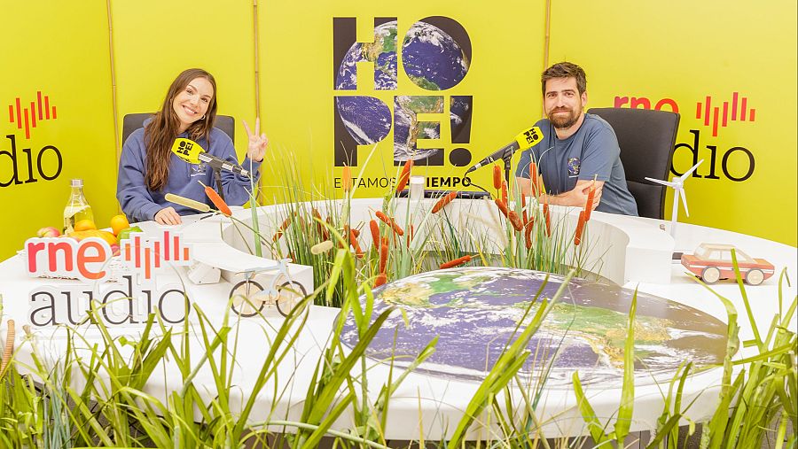 Dos presentadores de radio, uno con sudadera azul y otro con camiseta gris, sentados ante micrófonos en un estudio con decoración de temática ambiental.