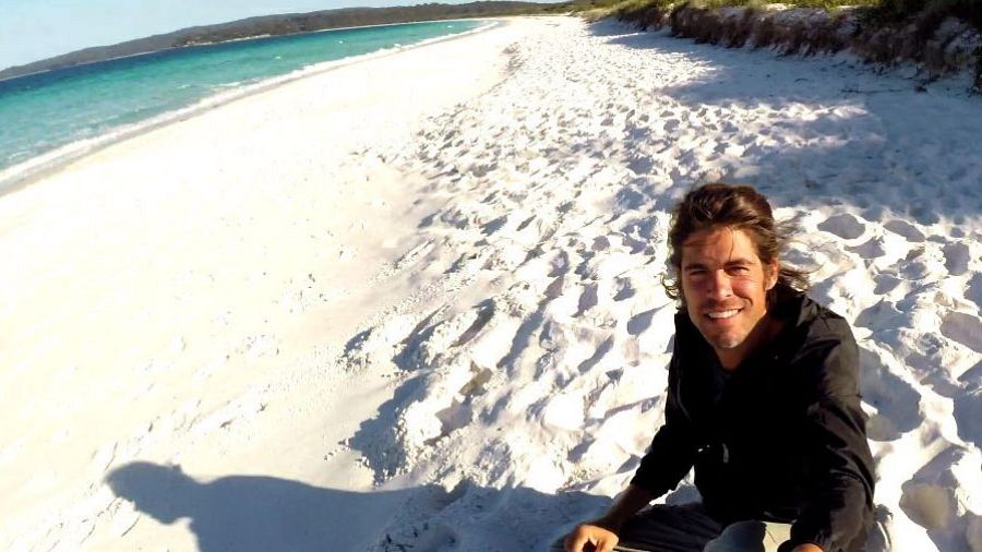 Selfie de un hombre en una playa paradisíaca de arena blanca y aguas turquesa, con vegetación de fondo.