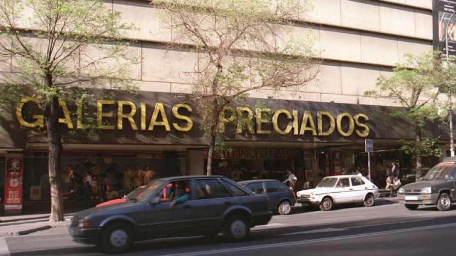 Fachada de Galerías Preciados con letrero dorado.  Árboles, coches y actividad visible a través de los escaparates.