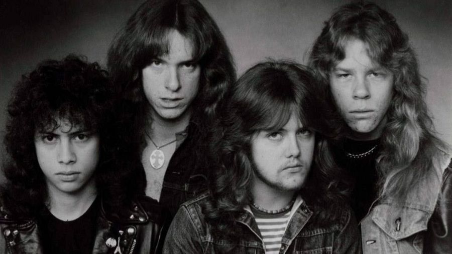 Metallica en blanco y negro, años 80.  James Hetfield, Lars Ulrich, Kirk Hammett y Cliff Burton con atuendos rockeros y melenas largas.