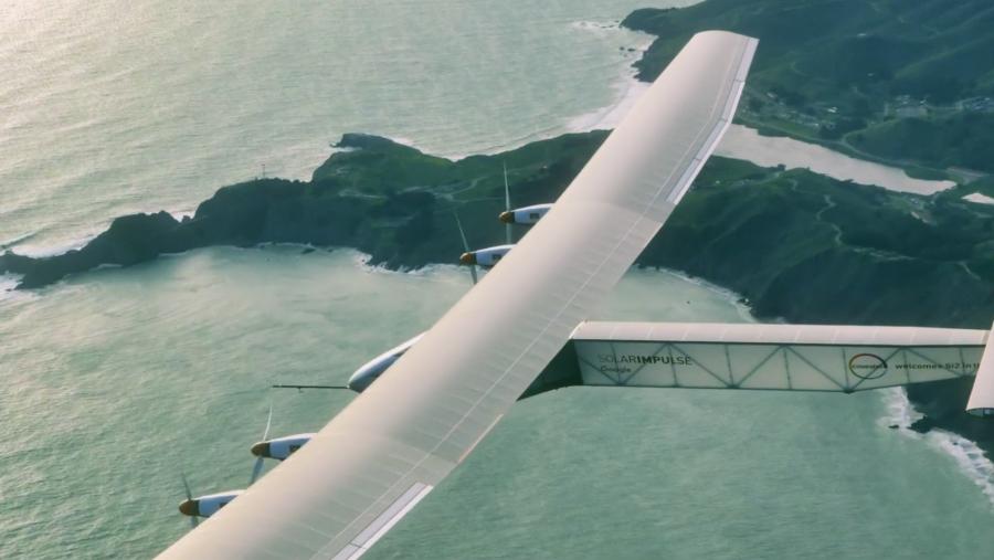 Vista parcial del avión Solar Impulse en vuelo sobre una costa.  Se observa el ala blanca con hélices, el logo de Schindler y el texto 'welcome's Ø12 in 17'.