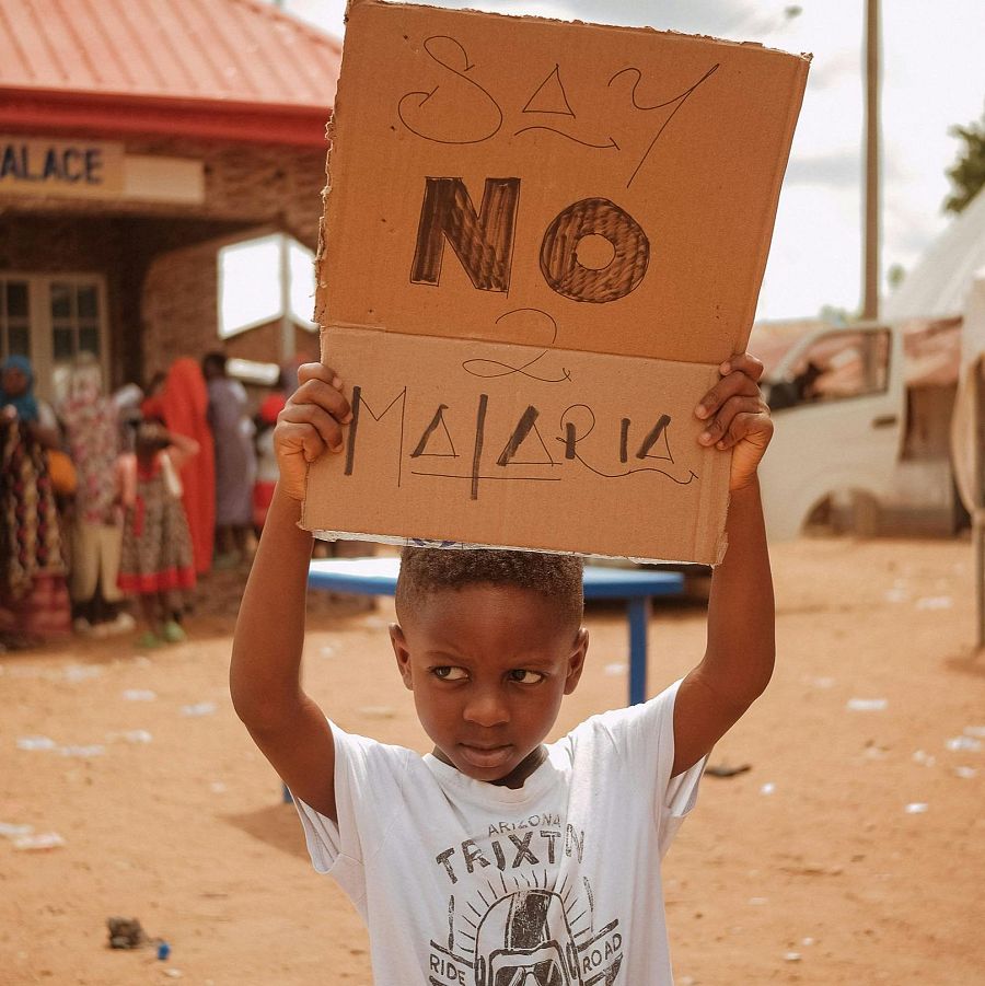 Niño con cartel "SAY NO MALARIA" mira a cámara.  Fondo con personas y posible edificio.  Imagen con mensaje contra la malaria.