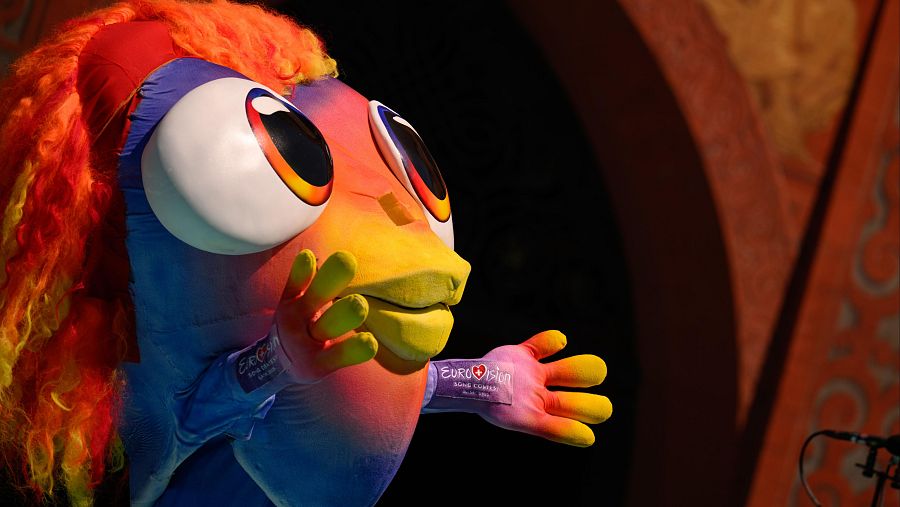Mascota colorida de Eurovisión con melena vibrante, guantes con logo y expresión amigable, sobre fondo oscuro.