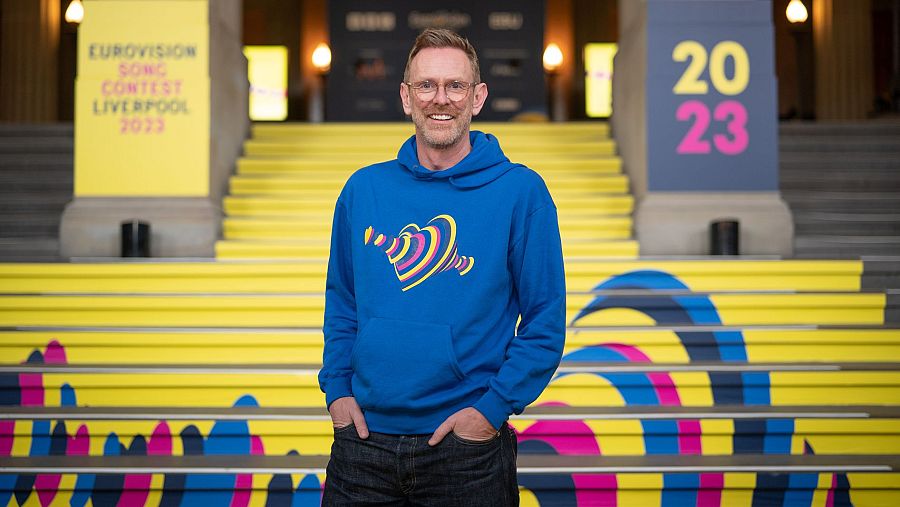 Director de Eurovisión 2025 con sudadera azul y logo del festival, frente a una escalera colorida cerca de carteles del concurso 2023 en Liverpool.
