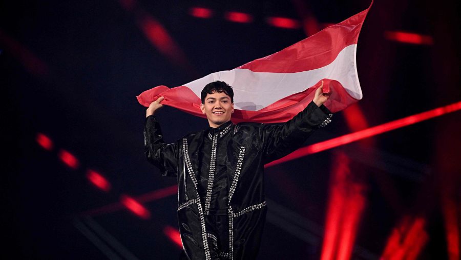 Representante de Austria en Eurovisión 2025 con traje negro y detalles plateados, porta la bandera austriaca durante el paseo de banderas en un escenario iluminado con luces rojas.