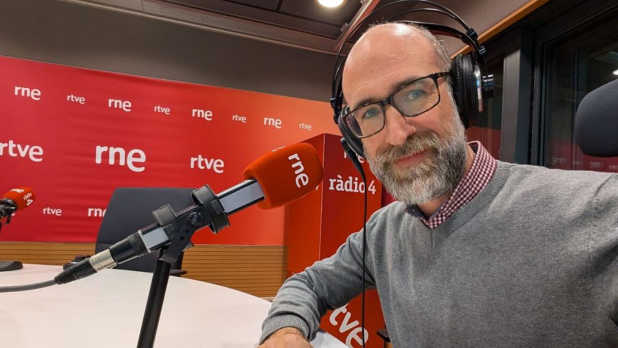 Periodista con barba canosa y gafas en estudio de radio RNE Barcelona, ante dos micrófonos; fondo rojo con logotipos RTVE y RNE.