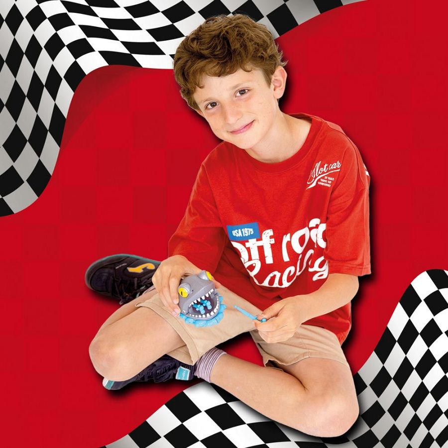 Niño sonriente con camiseta roja y juguete tiburón, sobre fondo rojo y bandera a cuadros.  Aspecto dinámico y enérgico.