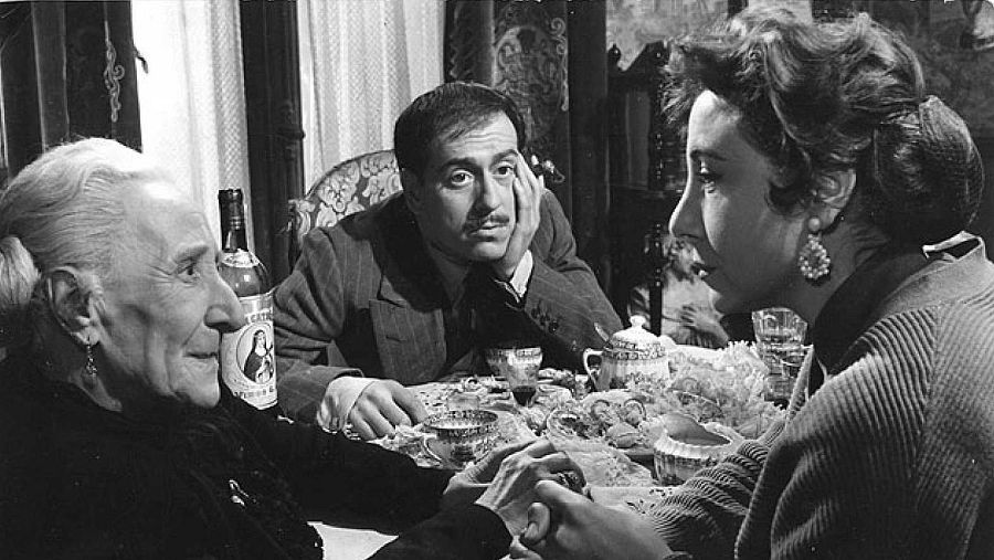 Fotografía en blanco y negro de una escena doméstica de 'El pisito' (1958), mostrando a José Luis López Vázquez y dos mujeres sentadas a una mesa con una botella de vino y tazas.