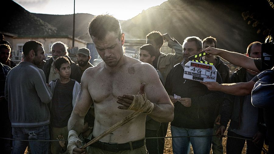 Matt Damon, vendado y con el torso descubierto, se prepara para una escena en un rodaje al aire libre.  El ambiente es polvoriento y se observa equipo de filmación.