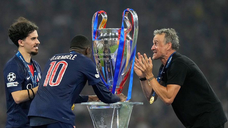 Jugadores de fútbol con medallas y su entrenador celebran, con el trofeo de la Champions League de fondo.  Se aprecia alegría y emoción.