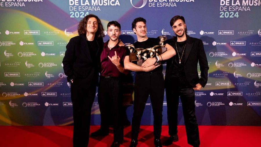 Los cuatro integrantes de Arde Bogotá posan con un trofeo en una ceremonia de premios sobre una alfombra roja, con un fondo azul y logos de patrocinadores.