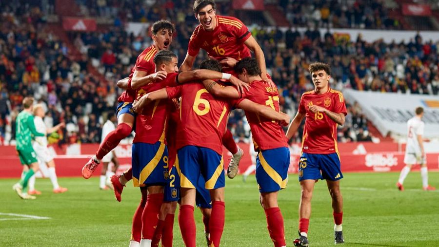 Celebración de gol de la selección española sub-21. Jugadores con camisetas rojas y azules se abrazan; se ven dorsales 16, 6, 15 y 9. Fondo con público y jugadores de otros equipos.