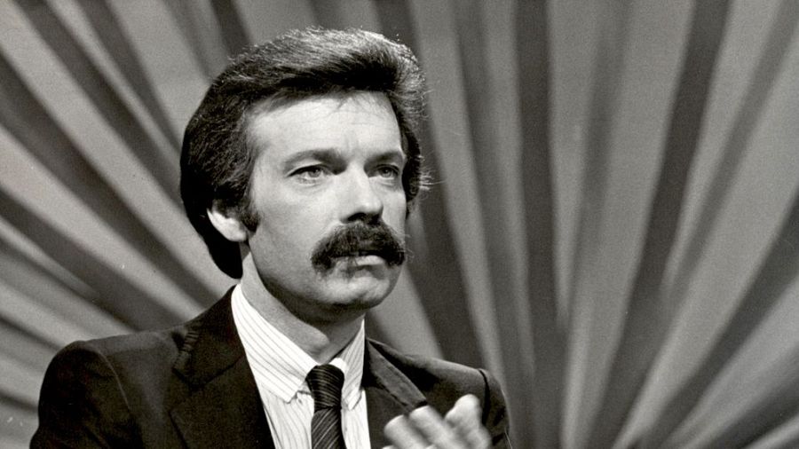 Presentador de televisión en blanco y negro, traje oscuro, camisa a rayas, fondo radial.  Imagen de estudio de televisión.