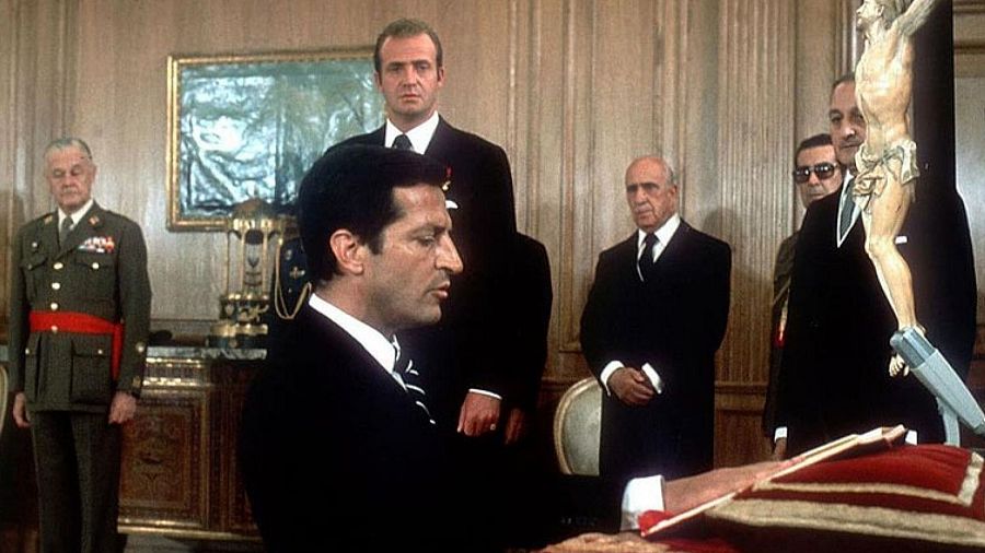 Adolfo Suárez juramento a la Constitución.  Presidente en traje oscuro, mano sobre libro rojo y dorado, en sala formal con otros hombres en traje y un militar.