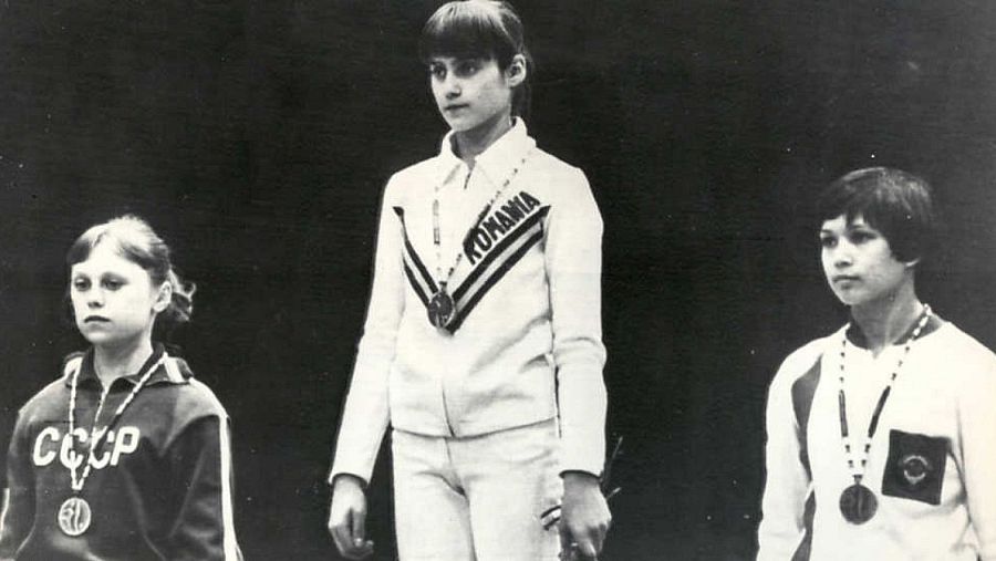 Tres gimnastas con medallas en un podio; una de ellas, con traje "ROMANIA", es Nadia Comaneci. Foto en blanco y negro.