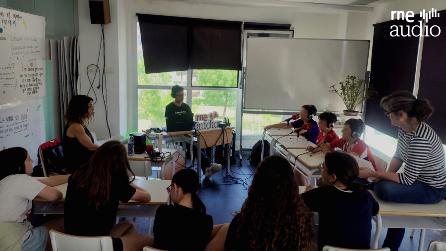 Emisión de radio escolar en un aula: alumnos con auriculares y equipo de audio, supervisados por una adulta. Se ve el logo de 'rne audio' y la pizarra con 'Sapiensantes colegio' y 'Radio'.