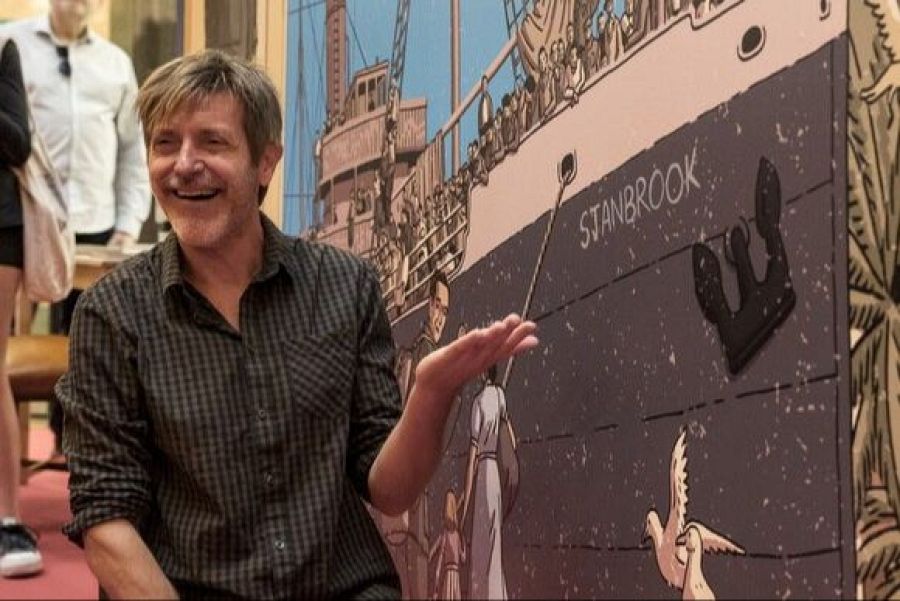 Inauguración de exposición de cómic español. Paco Roca gesticula ante ilustración de un barco lleno de gente, posiblemente representando un evento histórico.