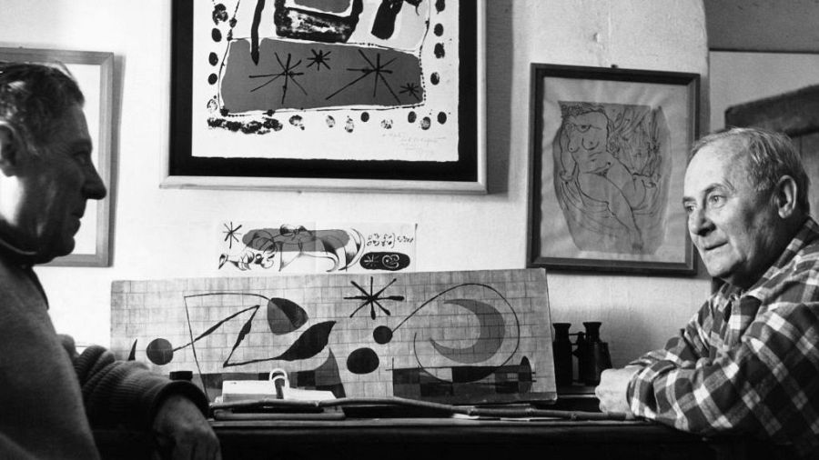 Dos hombres, sentados a una mesa, observan un mural abstracto con figuras geométricas y una luna creciente.  El ambiente sugiere un taller de trabajo artístico.