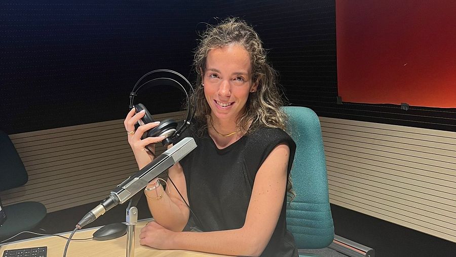 Mujer sonriente en estudio de radio con micrófono y auriculares. Viste camiseta negra. Fondo con paneles acústicos beige, oscuros y rojo anaranjado.