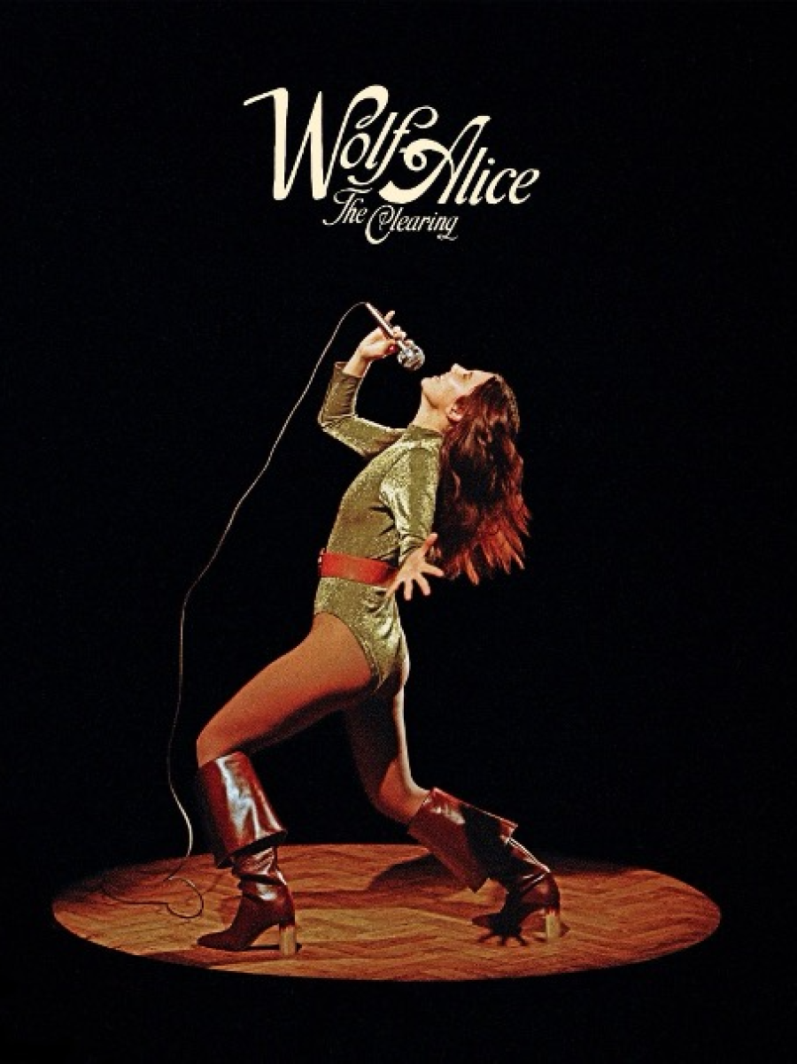 Cantante con mono dorado y botas marrones, micrófono en mano, sobre un escenario circular.  Texto "Wolf Alice" y "The Clearing" visibles.