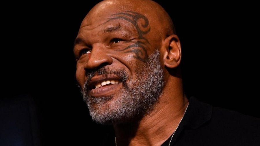 Primer plano de Mike Tyson con cabeza rapada, barba y bigote canosos, y un tatuaje tribal en la mejilla izquierda. Sonríe levemente con mirada hacia arriba y a la izquierda sobre fondo negro.
