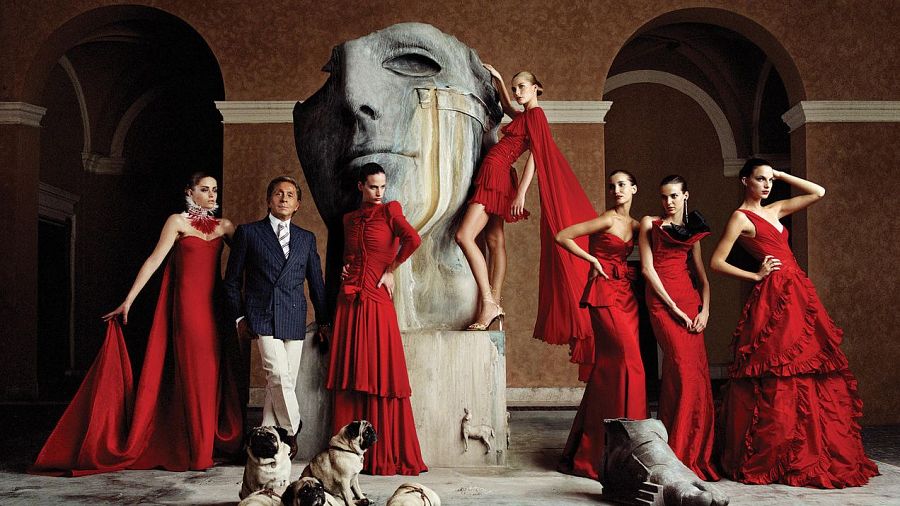Valentino Garavani, en traje azul marino, posa con modelos en vestidos rojos y sus carlinos en un entorno palaciego.