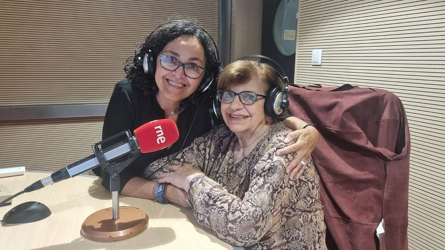 Dos mujeres sonrientes presentan un programa de radio en un estudio con micrófono RNE. Una lleva blusa negra y la otra una blusa estampada.